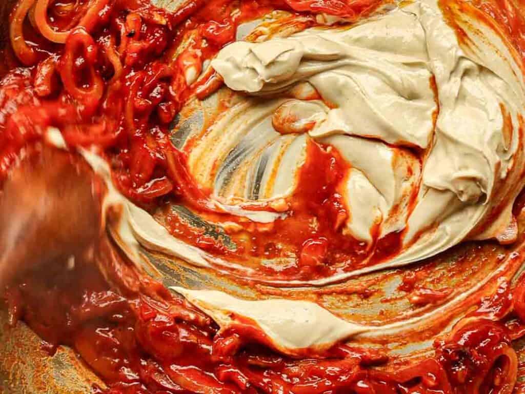 Close-up of a pan showing a rich tomato-based sauce being stirred with a creamy, light-colored ingredient, creating a swirling pattern. A wooden spoon is visible, partially mixing the vibrant red and cream elements.